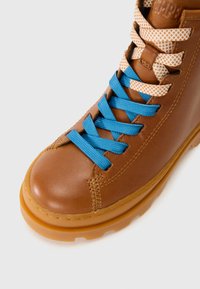 Botte en cuir brun avec un bout arrondi, ornée de lacets texturés bleu et crème, et d'une semelle en caoutchouc épaisse. Détails comprenant des coutures visibles.