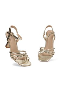 Tacones metálicos dorados con punta abierta, que presentan un diseño de tiras con un acento anudado y un tacón cuadrado para mayor estabilidad.