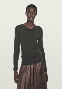 Maglia a maniche lunghe verde scuro con scollatura drappeggiata, abbinata a una gonna in satin marrone plissettata. Texture liscia, silhouette aderente e lucentezza sottile.