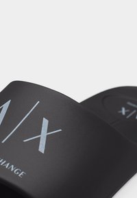 Μαύρο σάνδαλο με φαρδιά λουρί και το λευκό λογότυπο "A|X" τυπωμένο στο λουρί και στην επιφάνεια υποδημάτων, τοποθετημένο σε απλό φωτεινό φόντο.