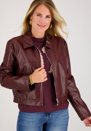 Vrouw met blond haar, gekleed in gouden oorringen, een bordeaux lederen ritssluiting jack, een bordeaux top met bloemmotief, spijkerbroek en gouden armbanden.