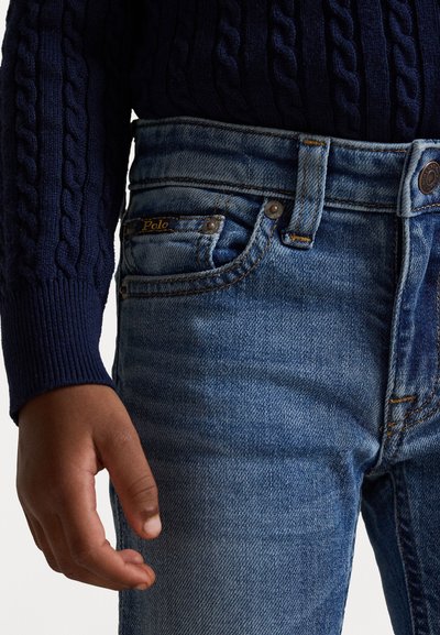 Jeans iz denima v srednje modri barvi z rahlim bledenjem, z zlatim šivom in oznako na sprednjem žepu. Kombinirani z temnomodrim puloverjem s pletenjem.