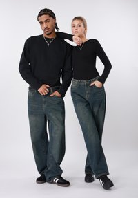 Δύο μοντέλα φορούν μαύρα πουκάμισα μακρυμάνικα. Φορούν μπλε τζιν παντελόνια με άνετη εφαρμογή. Τα τζιν έχουν τσέπες και ελαφρώς ξεθωριασμένη υφή.