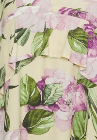 Um tecido com padrão floral apresenta grandes flores cor-de-rosa e verdes sobre um fundo amarelo pálido. Inclui um detalhe de barra com babados.