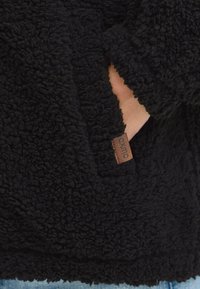 Pull en polaire noir avec une surface douce et texturée, poche latérale et étiquette en cuir marron portant le nom de la marque.