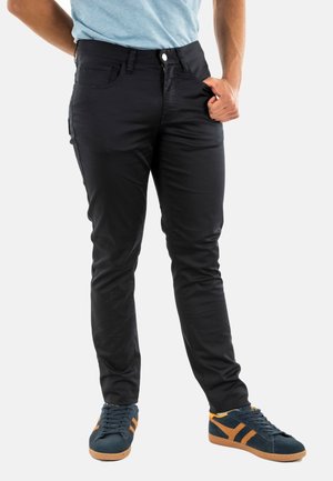 Pantalones negros de corte ajustado con textura suave, que cuentan con bolsillos delanteros y traseros, cremallera y cierre de botón. Combinados con zapatos azul oscuro.