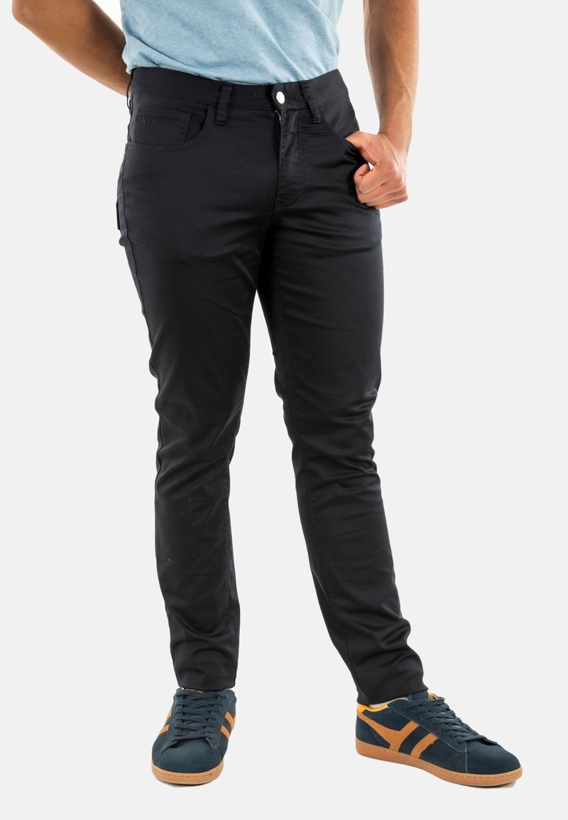 Pantalons noirs, coupe ajustée avec une texture lisse, contenant des poches avant et arrière, une fermeture éclair, et un bouton. Associés à des chaussures bleu foncé.