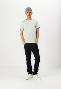 Lichtgroen T-shirt met korte mouwen, donkerblauwe jeans en witte sneakers. Pet is grijs met een logo. Model staat met handen in de zakken tegen een witte achtergrond.