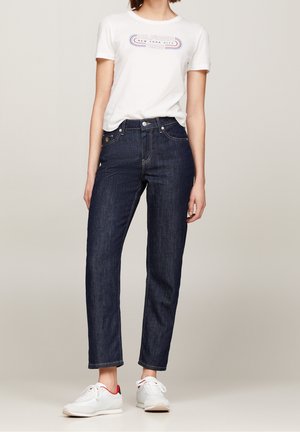Femme portant un T-shirt graphique blanc, un jean taille haute bleu foncé et des baskets blanches avec des accents rouges sur un fond neutre.
