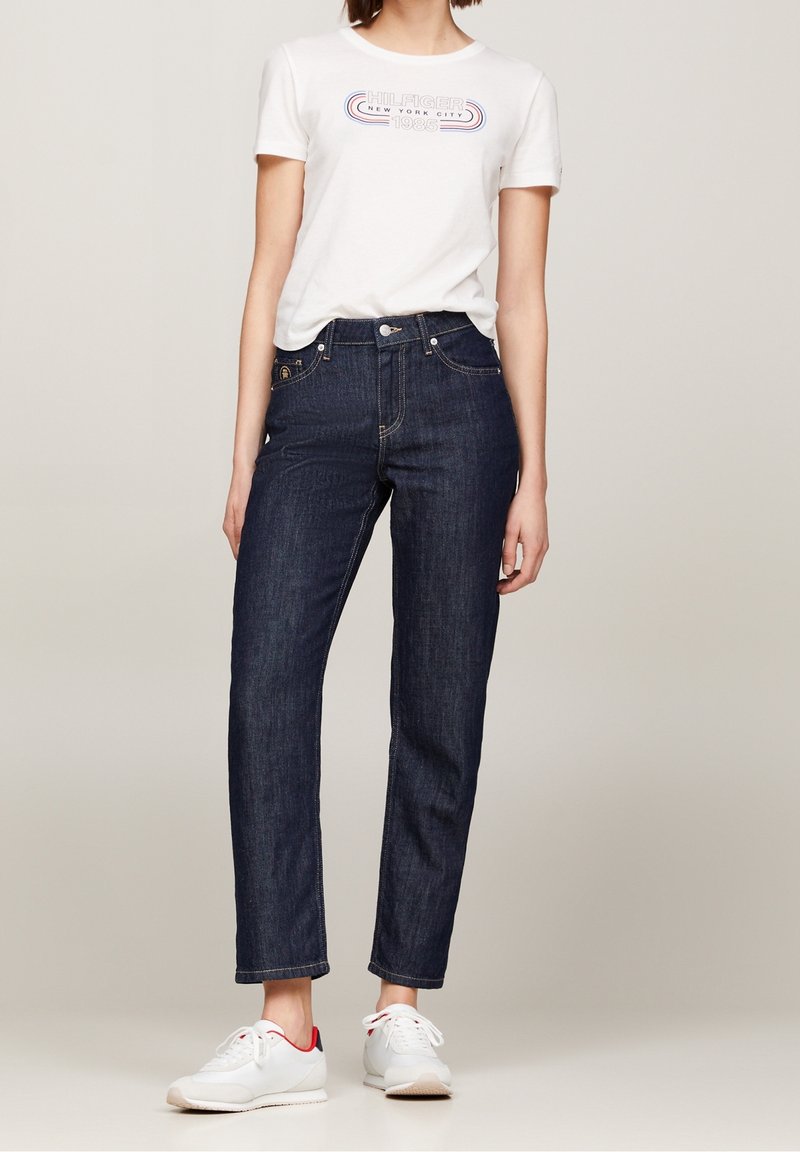 Femme portant un T-shirt graphique blanc, un jean taille haute bleu foncé et des baskets blanches avec des accents rouges sur un fond neutre.