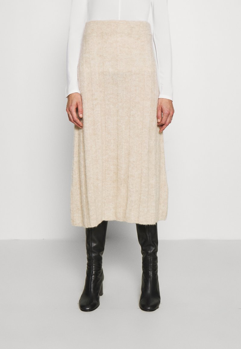 Saia midi de malha em bege claro, com textura canelada, cintura elástica larga e uma silhueta fluida e evasê. Combinada com botas pretas.