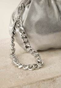 Silbermetallische Beuteltasche mit einem gerafften Design. Sie verfügt über einen massiven silbernen Kettenriemen und einen Verschluss, vor einem neutralen Hintergrund.