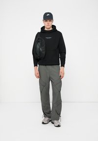 Čierna mikina s kapucňou a predným vreckom, šedé cargo nohavice s bočnými vreckami a šedé tenisky. Na ľavej strane je nosená čierna crossbody taška.