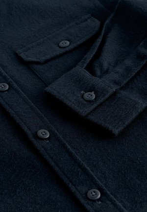 Schwarzes Wollhemd mit Knopfleiste, zwei Brusttaschen und strukturierter Stoffqualität. Verfügt über abgerundete Manschetten mit schwarzen Knöpfen.