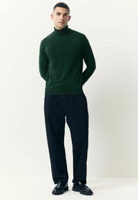 Pulover verde închis cu guler înalt, realizat dintr-un material moale, asortat cu pantaloni negri largi și pantofi negri, creând o ținută lejeră.