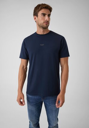 Mand med kort brunt hår iført en marineblå Guess t-shirt og blå jeans, der står foran en ensfarvet lys grå baggrund.