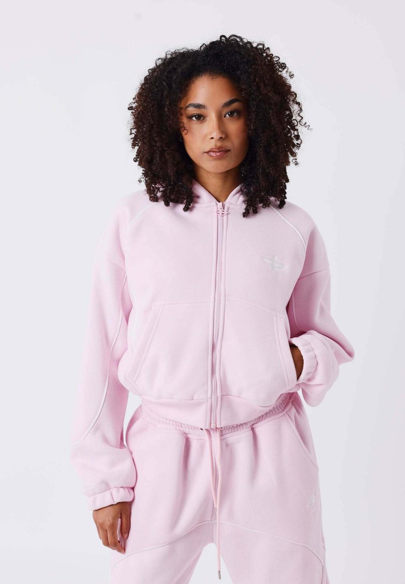 Sweat à capuche rose zippé en tissu doux, avec une capuche, des poches avant et des poignets côtelés. Associé à un pantalon assorti pour un look harmonieux.