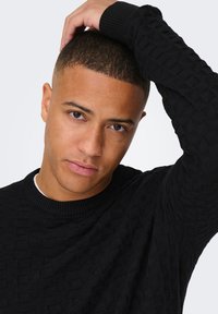 Schwarzer gestrickter Pullover mit einem strukturierten Rautenmuster, Rundhalsausschnitt und gerippten Bündchen. Das Model posiert mit einer Hand auf dem Kopf.
