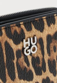 Novčanik s leopard uzorkom koji sadrži teksturiranu smeđu i crnu površinu, ukrašen srebrnim "HUGO" logotipom i zatvaranjem na munju na vrhu.
