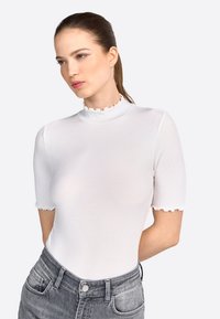 Hvid ribbet bodysuit med korte ærmer, der har en flæset høj hals og en tætsiddende design. Kombineret med højtaljede grå jeans.