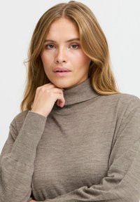 Pull à col roulé beige en tissu tricoté doux. Il présente des manches longues ajustées et un col haut. Texture subtile avec une finition lisse.