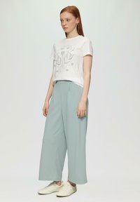 Witte katoenen t-shirt met groen grafisch ontwerp, gecombineerd met lichtblauwe wijde broek, aangevuld met beige sneakers.