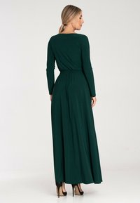 Długa zielona sukienka maxi z długim rękawem, dopasowaną talią, zwiewnym dołem i gładkiego materiału. Noszona z czarnymi szpilkami. Widok z tyłu.