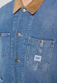 Chaqueta de denim en azul claro con un cuello de pana marrón, que cuenta con dos bolsillos delanteros y botones dorados con el logo de Lee.