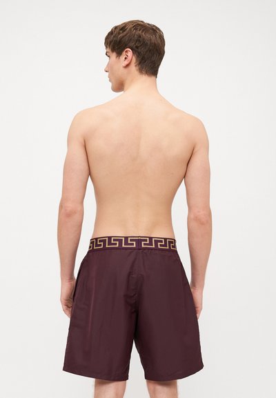 Κοτλέ μαγιό σε μπορντό χρώμα με χρυσό σχέδιο ελληνικού κλειδιού στη ζώνη. Λείος υφή, μήκος μέχρι το μέσο του μηρού, και ελαστική μέση για άνεση.