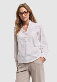 Lekka, biała bluzka w pionowe beżowe paski, z kołnierzykiem mandarynkowym, guzikiem i koronkowymi detalami na klatce piersiowej. Połączona z beżowymi spodniami.