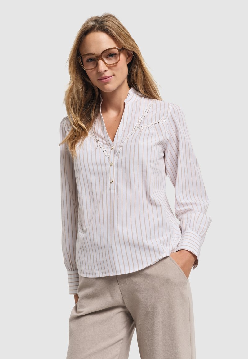 Lekka, biała bluzka w pionowe beżowe paski, z kołnierzykiem mandarynkowym, guzikiem i koronkowymi detalami na klatce piersiowej. Połączona z beżowymi spodniami.