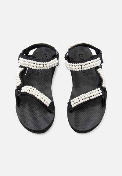 Μαύρα σανδάλια με υφασμένα πέλματα και πολλαπλούς ιμάντες διακοσμημένους με σειρές λευκών ψεύτικων μαργαριταριών σε λευκό φόντο.