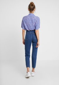 Žena s vlasy v drdolu, která nosí náušnice ve tvaru kruhu, krátký rukáv s fialovým vzorem, vysoké modré džíny a bílé tenisky, stojí zády.