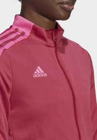Chaqueta atlética roja con detalles en rosa, que presenta un cuello alto, cierre de cremallera y tres franjas rosas en los hombros. Logotipo de Adidas visible.