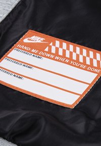 Tissu noir avec une étiquette orange présentant le logo "Nike", le texte "PASSE-MOI QUAND TU AS TERMINÉ" et des espaces pour des noms. Bords texturés.