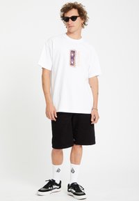 Hvid t-shirt med kort ærme og et farverigt grafisk design på brystet, parret med sorte shorts, hvide sokker og sorte sneakers.