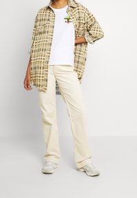 Karierte Hemd in Beige- und Brauntönen über einem weißen T-Shirt, kombiniert mit off-white Cordhosen und hellgrauen Sneakers.