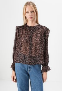 Blusa nera trasparente con motivo floreale, colletto arricciato, chiusura a bottoni frontale e maniche a campana; indossata con jeans azzurri.