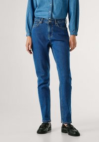 Modré džíny z denimu s přiléhavým střihem, pětikorunovou kapsou a malým knoflíkovým zapínáním, doplněné černými botami. Hladká textura, bez viditelných vzorů.