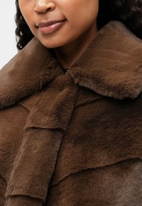 Brązowa kurtka z faux futra z szerokim kołnierzem, miękką teksturą i warstwowym designem z widocznymi detalami przeszycia.