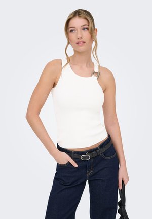 Donna con top bianco senza maniche con dettaglio ad anello in metallo e jeans blu scuro, che tiene una borsa nera in una mano.