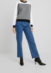 Felpa a maniche lunghe con motivo in bianco e nero, abbinata a jeans a gamba larga in denim blu e stivali neri alla caviglia, in piedi davanti a uno sfondo neutro.