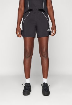 Personne portant un short de sport noir, des chaussures de course noires, des chaussettes blanches et un haut noir, debout devant un fond uni.