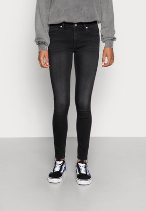 Persoon die zwarte skinny jeans, een grijs shirt met lange mouwen en zwart-blauwe sneakers draagt, lopend tegen een effen lichte achtergrond.