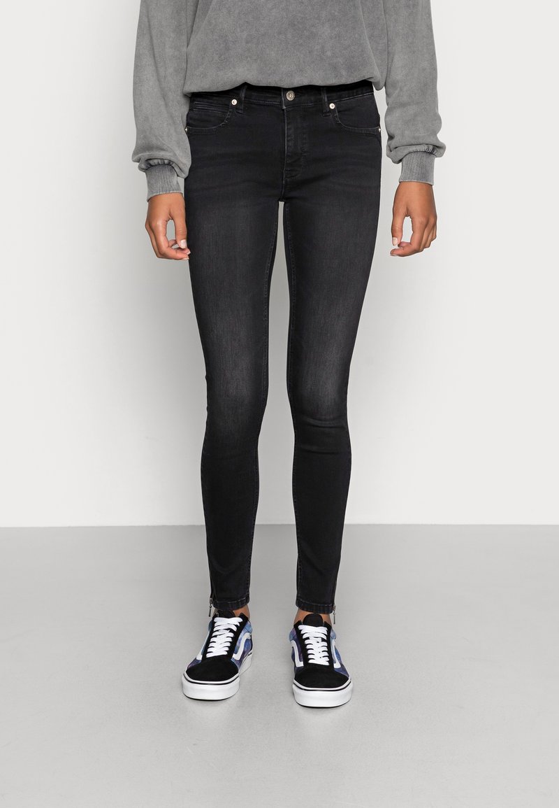 Personne portant un jean skinny noir, un haut gris à manches longues, et des baskets noires et bleues, debout devant un fond clair uni.