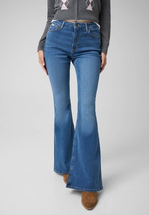 Kvinde iført højtaljede blå vidde jeans, grå kort sweatshirt med lyserød rombemønster og brune spidse støvler, stående foran en ensfarvet baggrund.