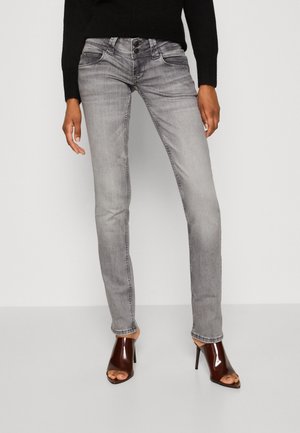 Persoon draagt strakke lichtgrijze spijkerbroek met drievoudige knoopdetail en donkerbruine open-toe hoge hakken, staand tegen een effen achtergrond.