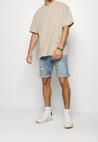 Béžové oversized tričko, světle modré roztrhané denimové šortky s ohrnutými lemy, doplněné bílými teniskami a kotníkovými ponožkami.