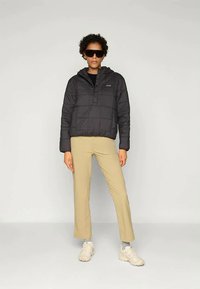 Person med svart quilted jakke, beige bukser, hvite sko, grå sokker og store svarte solbriller, står mot en enkel hvit bakgrunn.