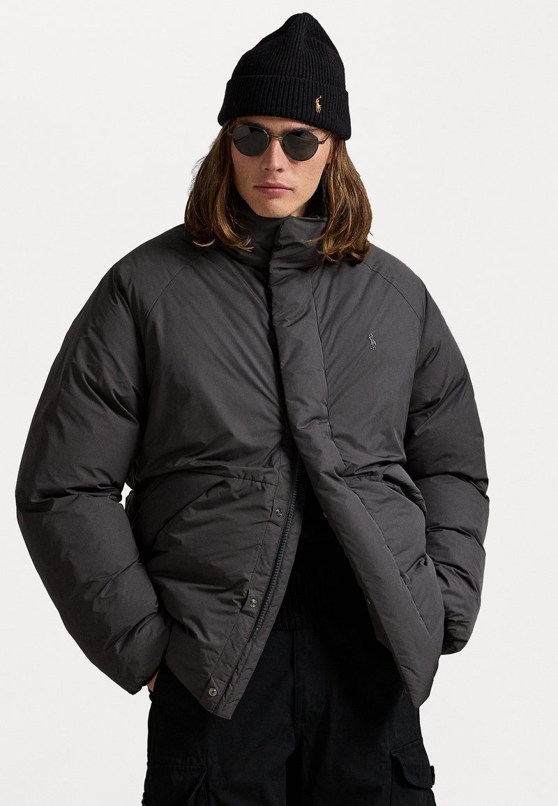 Unge person iført sort beanie, runde solbriller, mørk quilted jakke og sorte cargo-bukser med hænderne i lommerne mod en hvid baggrund.